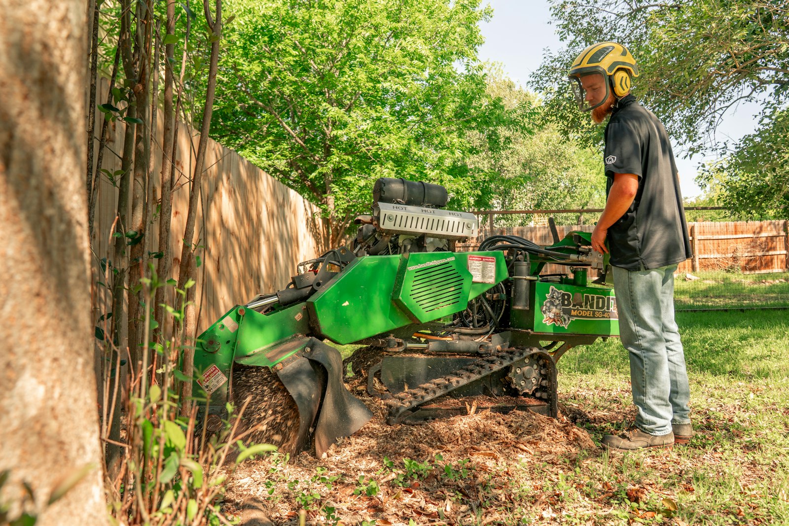 Operator grinding a tree stump with a tracked grinder in a residential backyard beside a privacy fence
